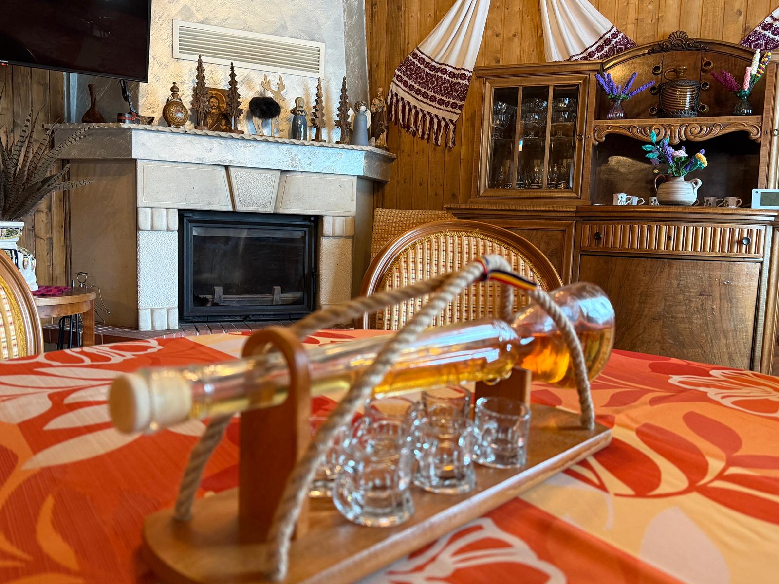 Un living de cabană cu decor tradițional, prevăzut cu șemineu, mobilier vechi din lemn și un suport decorativ cu sticlă de băutură și pahare mici pe masă.