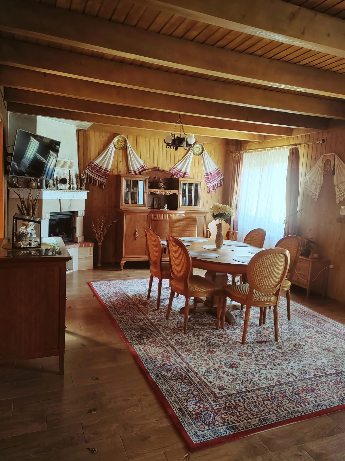 Living spațios, decorat în stil tradițional, cu o masă mare de dining în centru, șemineu, mobilier din lemn masiv și un covor cu modele clasice.